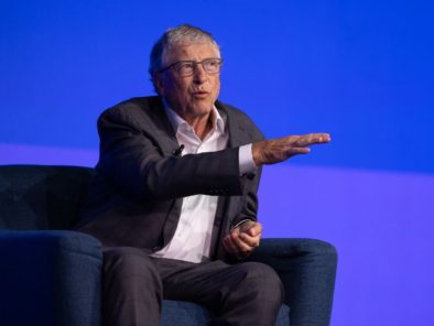 An older man in a suit and glasses is speaking on stage, gesturing with one hand, against a blue background.