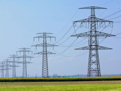 A row of tall metal electricity pylons stretches across a field under a clear blue sky.