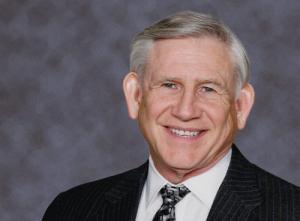 Smiling older man with gray hair wearing a black pinstripe suit and patterned tie, set against a neutral gray background.