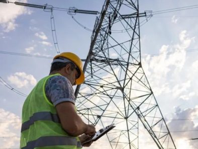 A worker in a yellow hard hat and vest examines a clipboard near a large power line tower, set against a partly cloudy sky.