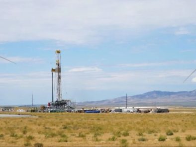 Wind turbines flank an oil rig in a vast desert landscape under a partly cloudy sky.