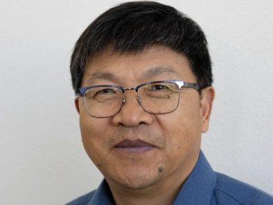 A man with short black hair and glasses is wearing a blue shirt against a plain background.