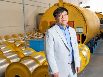 A man in a light suit stands in a factory, surrounded by large yellow machinery and metal coils.