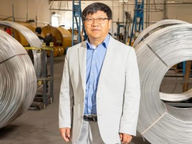 A person in a light suit stands in a warehouse with large coils of wire in the background.