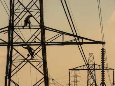 Two workers climb a large electric transmission tower, with several other towers visible in the background against a cloudy sky.