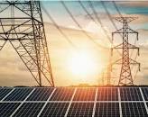 Solar panels in the foreground with transmission towers silhouetted against a setting sun in the background.