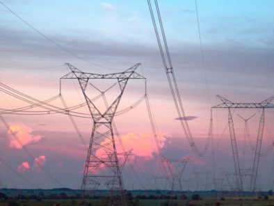 Power lines stretch across a rural landscape during sunset, with a colorful sky of pink and blue hues in the background.