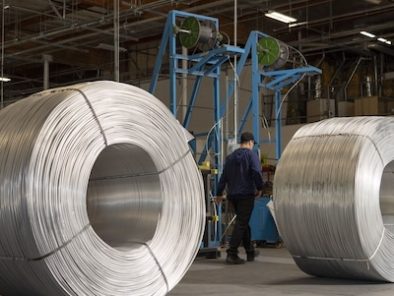 Large coils of metal wire are placed in a spacious industrial facility. A worker in a blue uniform walks past the coils. Machinery and overhead pipes are visible in the background.