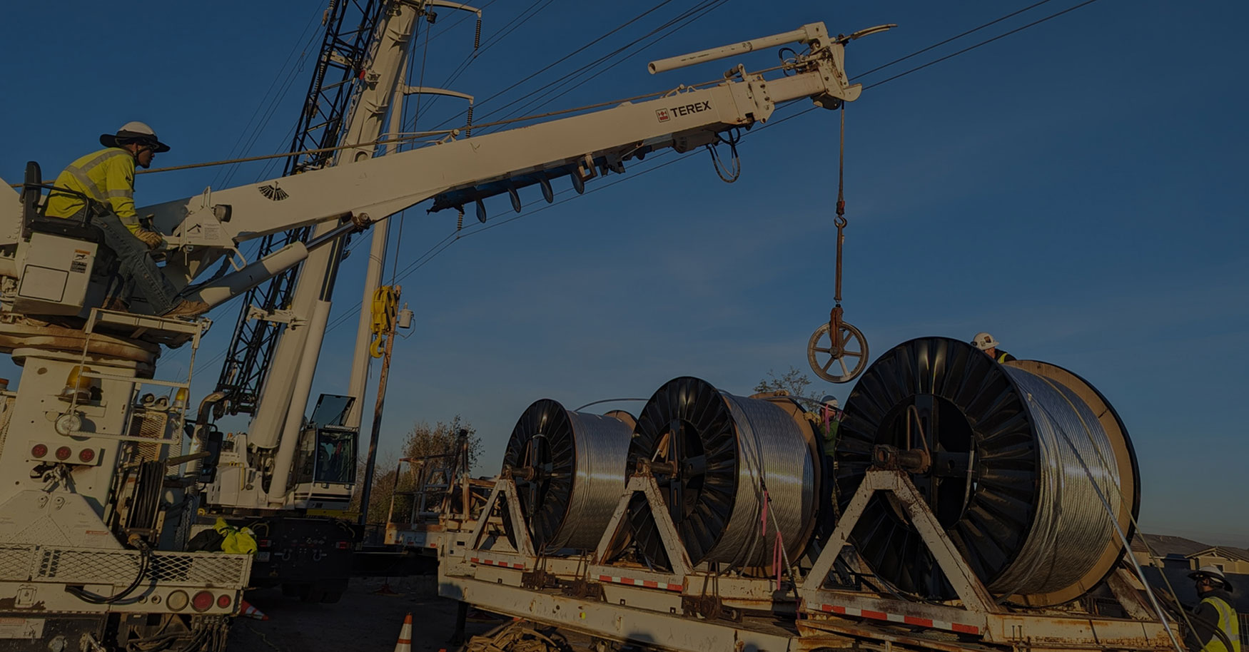 Construction worker operates a crane, lifting large cable spools from a flatbed truck on a clear day.