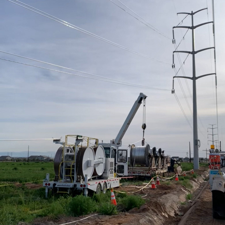 Utility workers operate machinery with large spools and a crane near power lines in a grassy area, with safety cones along the path.