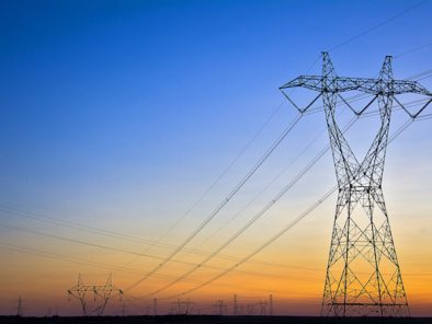 High-voltage power lines and towers stretch across a landscape at sunset, silhouetted against a clear sky.