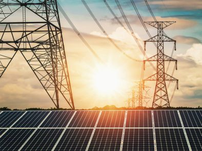 Solar panels in the foreground with electrical transmission towers under a sunset sky in the background.