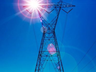 A power line tower against a clear blue sky with the sun shining brightly, creating lens flares.