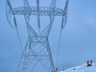 Two workers in orange stand near a large electrical tower in a snowy landscape, with overcast skies in the background.