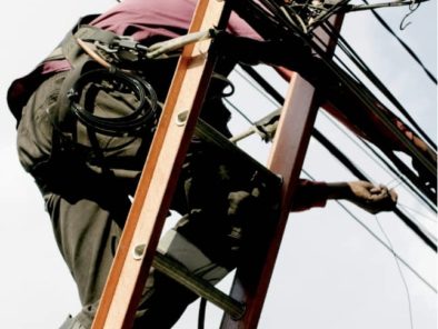 A person on a ladder works with multiple cables, wearing safety gear against a cloudy sky.