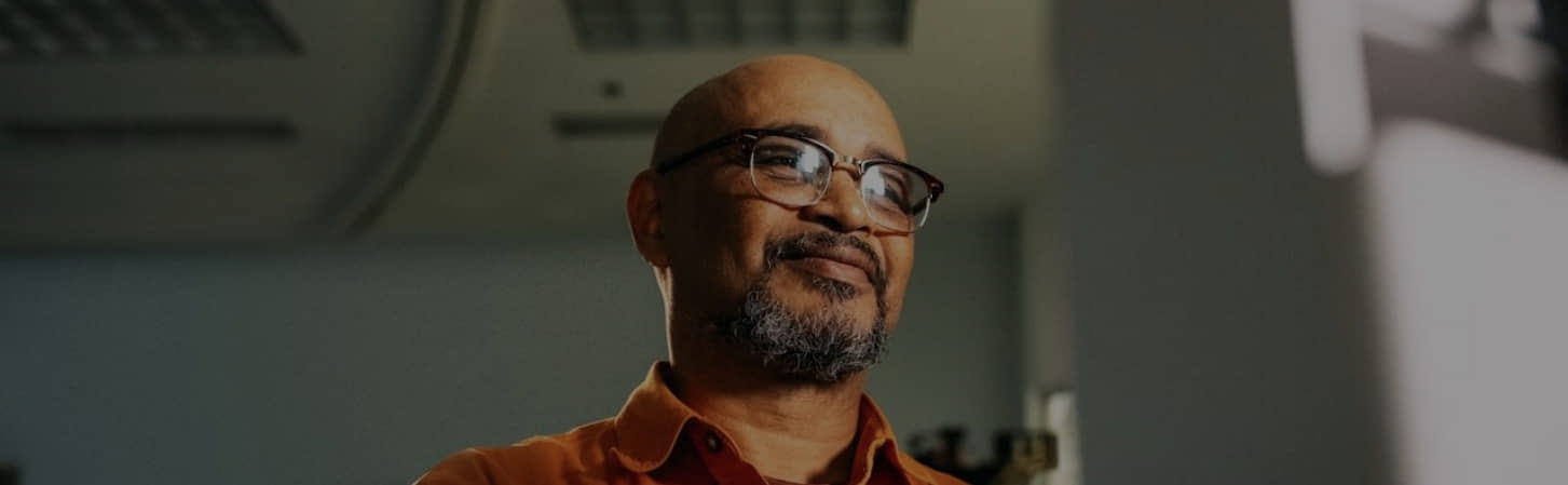 A man with glasses and a beard smiles softly, wearing an orange shirt, in a dimly lit room.