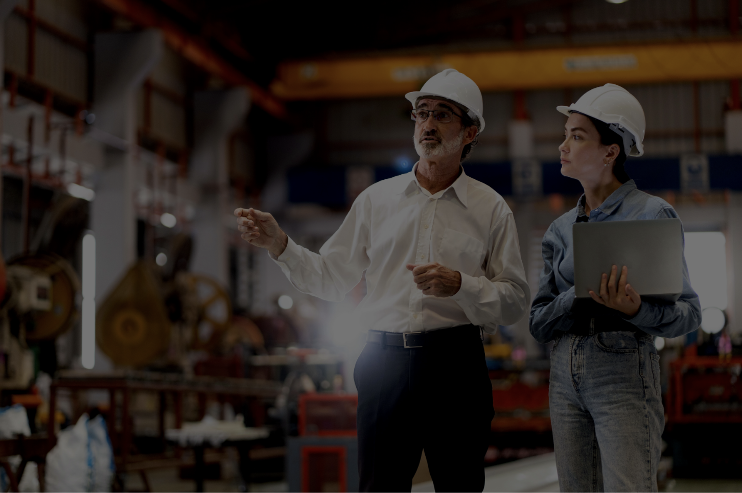 Two individuals in safety helmets are in an industrial setting. One holds a laptop, while the other gestures, possibly discussing something related to the machinery in the background.