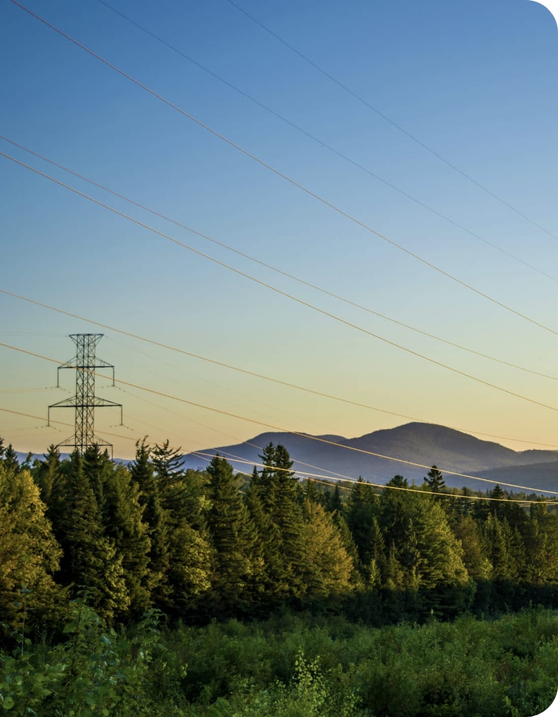 A transmission tower and power lines stand above a forested landscape with mountains in the background under a clear blue sky at sunset.