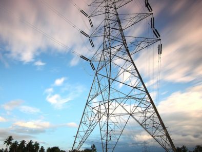 A tall electricity pylon stands in a green field under a cloudy sky, with power lines extending into the distance.