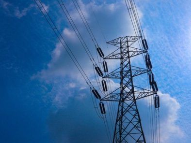 A metal electricity pylon with multiple power lines set against a blue sky with some clouds and a bright sun.