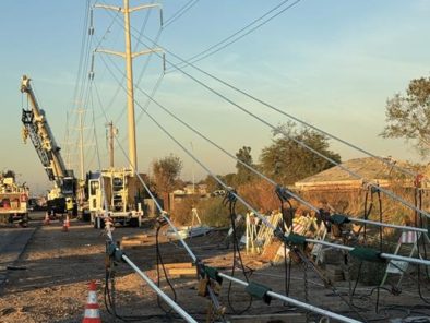 Construction site with utility trucks and a crane working on power lines. Several cables are laid out on the ground near power poles.