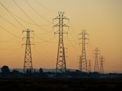 Power lines with metal pylons stretch across a landscape at sunset, silhouetted against an orange sky.