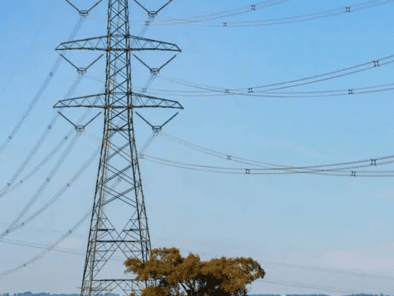 A tall electrical transmission tower stands on a grassy hill with several power lines extending from it under a clear blue sky. A tree is nearby.