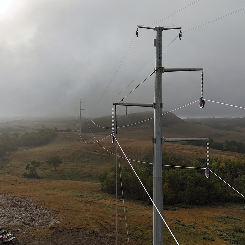 Power lines stretch across a foggy landscape with hills and vegetation below, under an overcast sky.