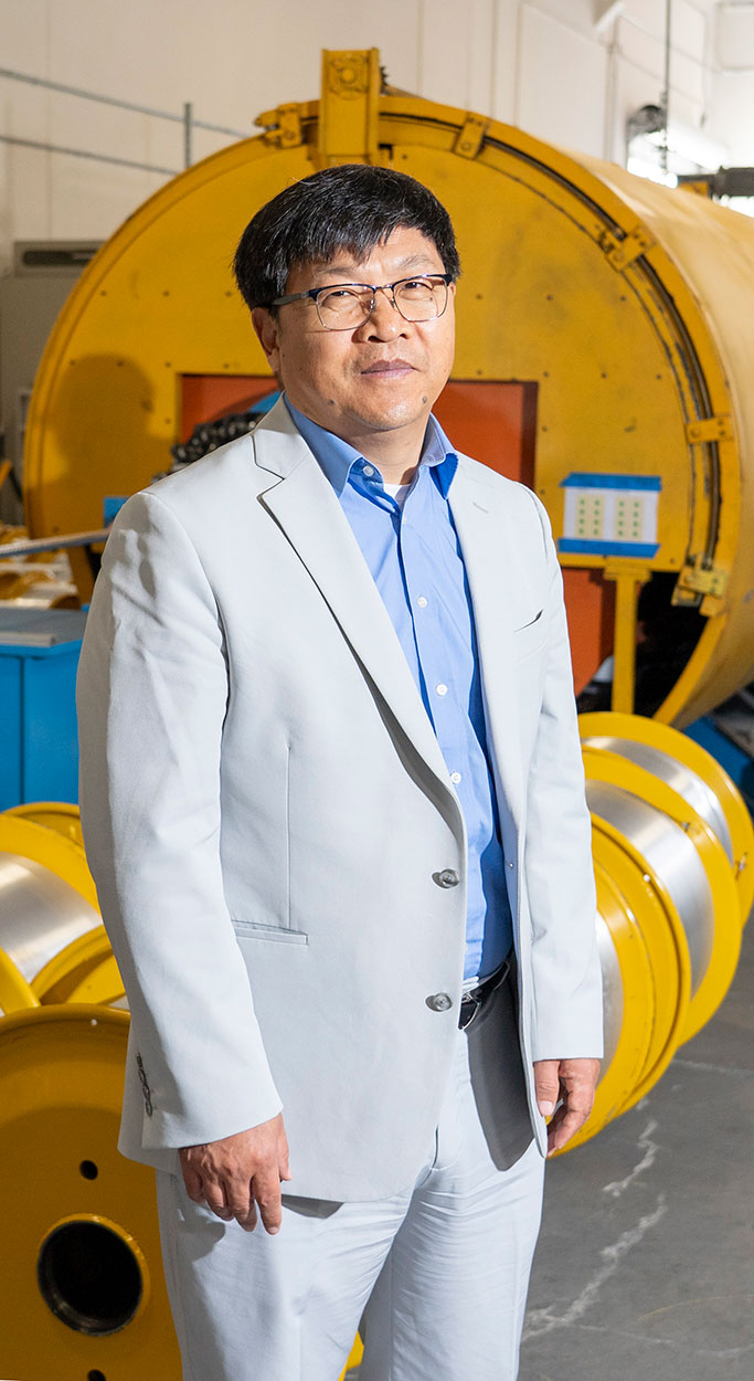 A man in a light suit stands in front of large yellow industrial equipment in a warehouse.