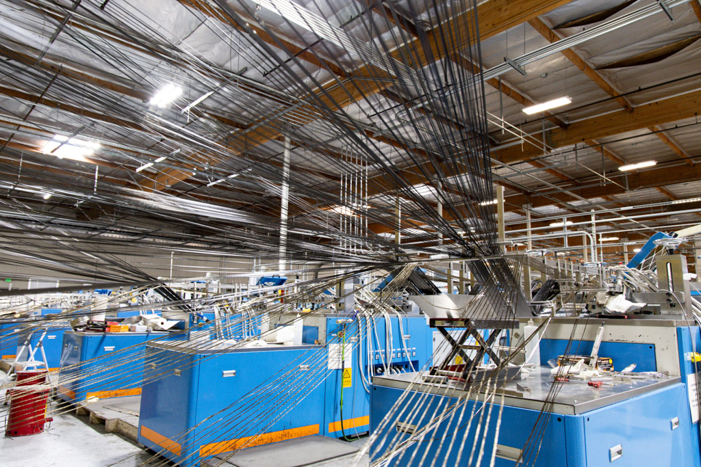 A factory interior with blue industrial machines, metal rods, and numerous cables or wires suspended above, with fluorescent lights on the ceiling.