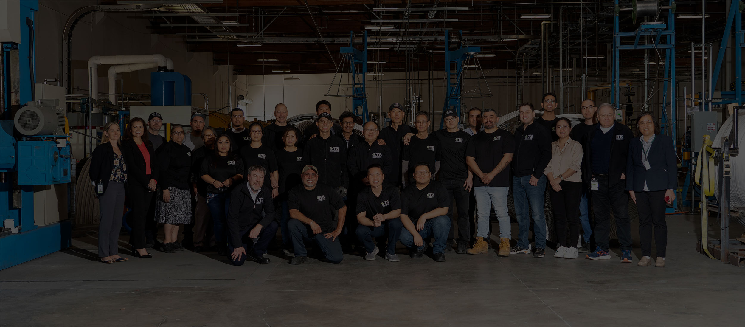 A diverse group of people wearing black shirts pose together in an industrial setting.