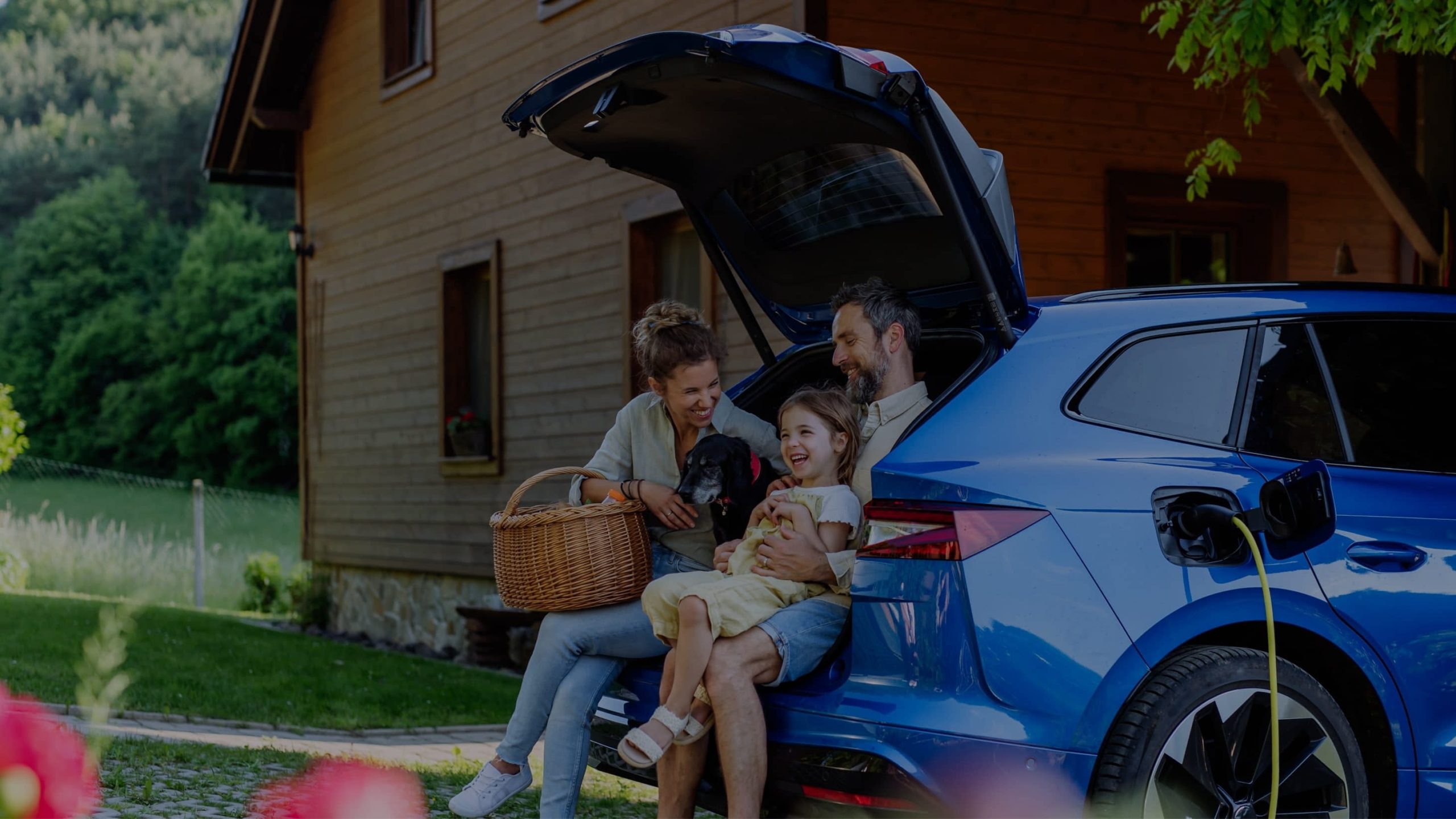 A family sits on the open trunk of a blue electric car parked near a wooden house, enjoying time together. The trunk contains a picnic basket and a dog.