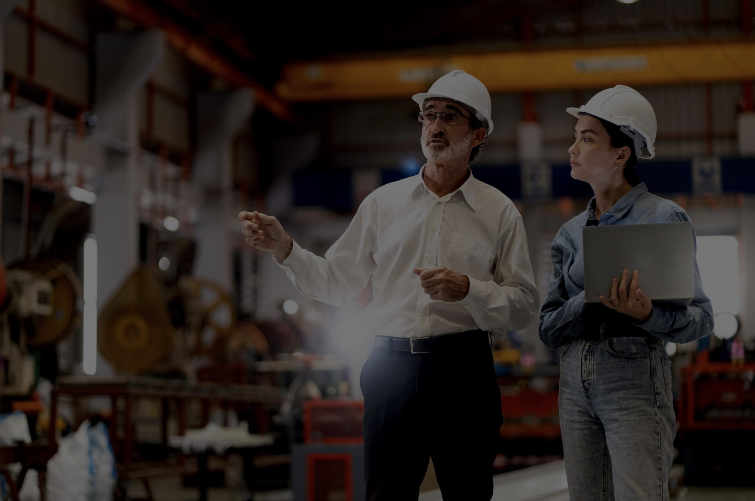 Two people in hard hats stand in a factory setting; one gestures while the other holds a laptop.