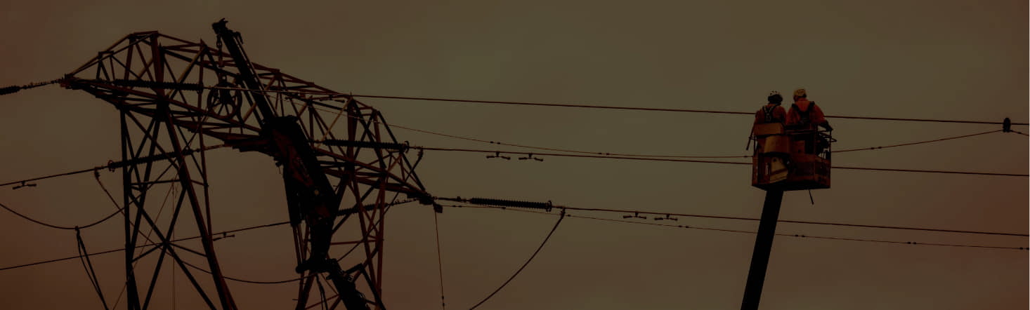 Two workers in a lift perform maintenance on a high-voltage power line near a large transmission tower, under a cloudy sky.