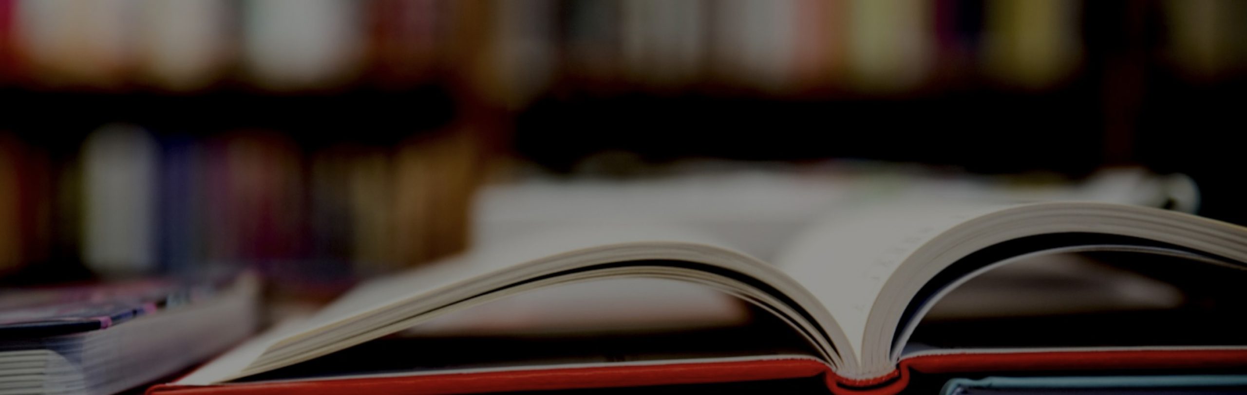 An open book with a red cover lies on a table, with a blurred background of shelves filled with books.