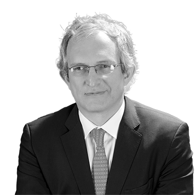 A man in a suit and tie with glasses is seated against a dark background.