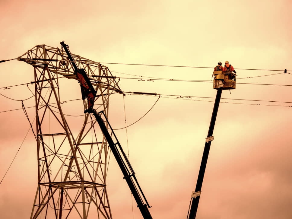 Workers in a lift repair electrical wires on a transmission tower against a cloudy sky.