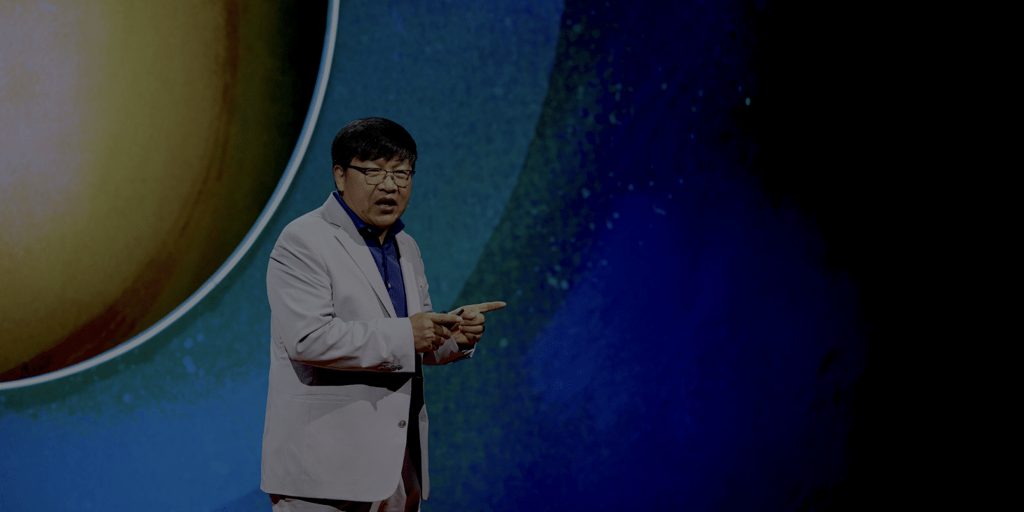 A person in a light suit speaks on stage with a vivid celestial-themed background.