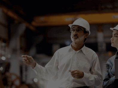 Two people wearing hard hats stand in an industrial setting; one gestures while talking, and the other holds a laptop, listening attentively.