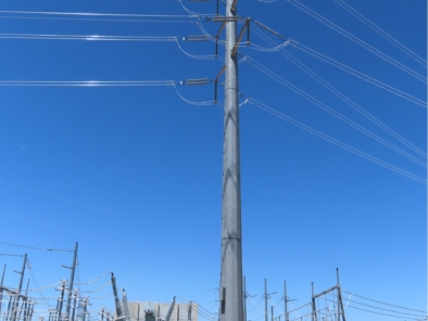 A tall electrical transmission tower stands in a power substation under a clear blue sky, with multiple power lines extending outward.