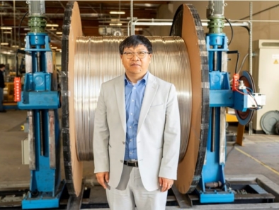 A man in a light gray suit stands in front of large industrial wire spools inside a factory setting.